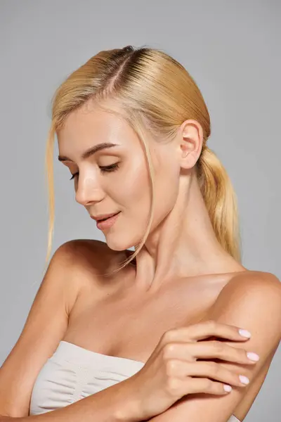 Chica con el pelo rubio y la piel clara mira hacia abajo y se abraza contra en fondo gris - foto de stock