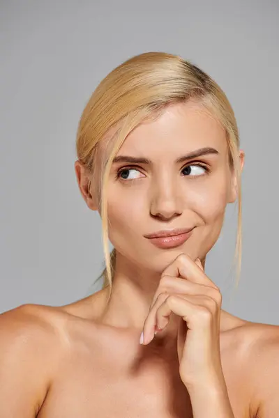 Mujer joven con ojos grises y cabello rubio pensando y mirando al lado en fondo gris - foto de stock