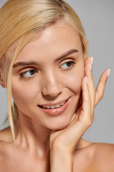 Beauty girl with blonde hair looking to side and hugs face with hand against in gray background — Stock Photo