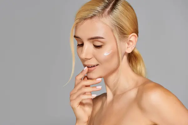 Alluring girl in her 20s  with cream on her cheek touching her lips against grey background — Stock Photo
