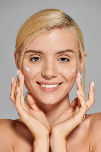Portrait smiling young woman with hands around her face and cream on cheeks against gray background — Stock Photo