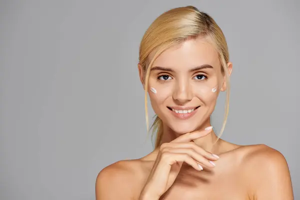 Alluring girl in her 20s with cream on cheeks touching her chin against gray background — Stock Photo