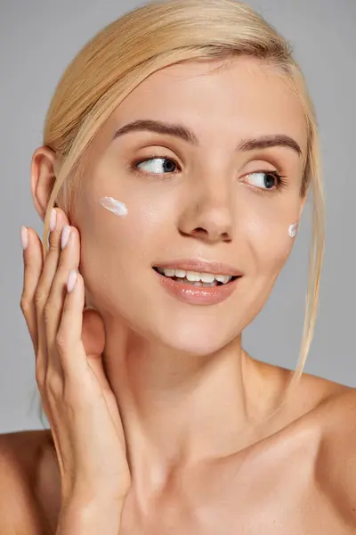 Beauty girl in her 20s applying cream on her cheeks and looking to side against grey background — Stock Photo