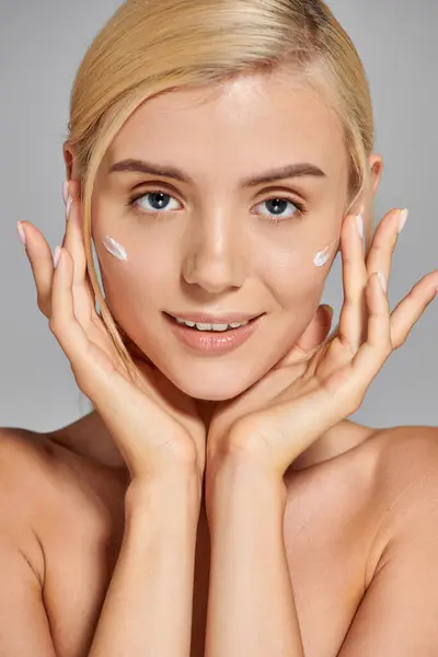 Charming young woman with hands around her face and cream on cheeks against gray background — Stock Photo