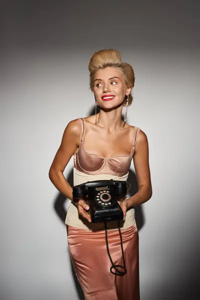 Mujer joven con estilo posando con teléfono retro y mirando hacia un lado contra el fondo gris - foto de stock