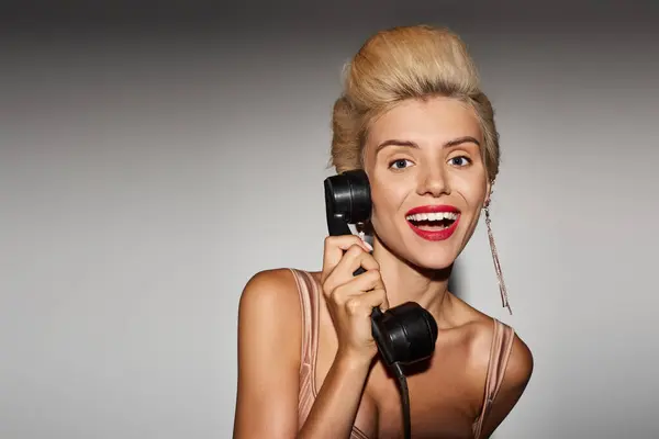 Chica con estilo en los años 20 con el pelo rubio recoge el teléfono retro contra el fondo gris - foto de stock