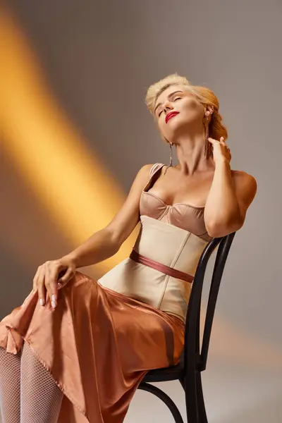 Задоволена блондинка молода жінка в корсетному платті позує зі стільцем на сірому фоні — стокове фото