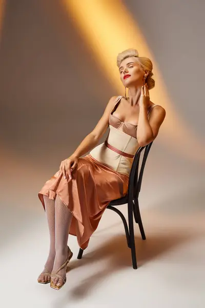 Chica satisfecha en 20s con pelo rubio en traje de corsé posando con silla sobre fondo gris - foto de stock