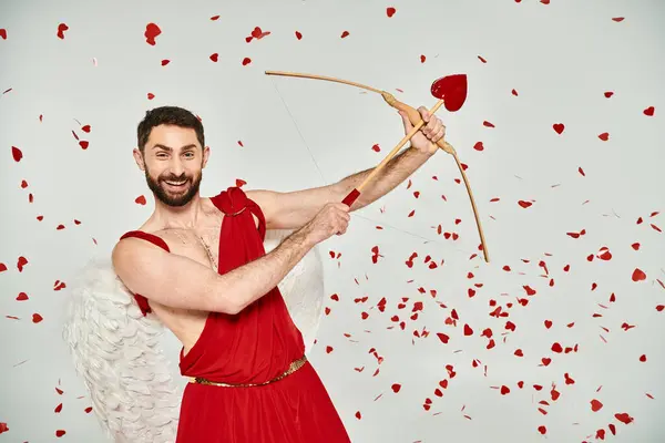 Excité homme cupide barbu tir à l'arc sous confettis en forme de coeur sur gris, st fête de la Saint-Valentin — Photo de stock