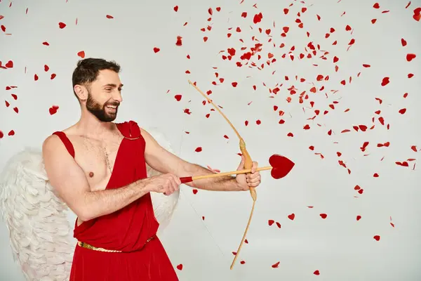 Sonriente hombre Cupido barbudo arqueando con flecha en forma de corazón bajo confeti rojo, día de San Valentín - foto de stock