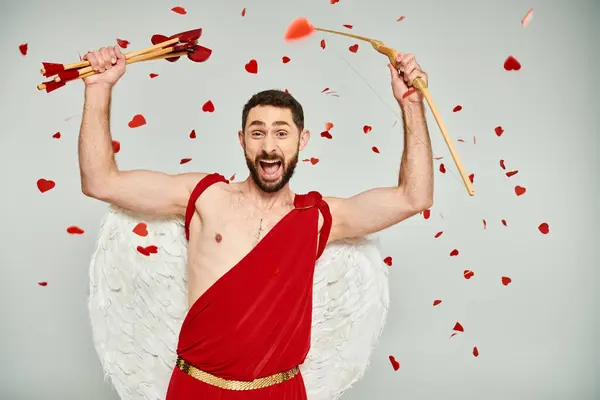 Hombre barbudo vestido de Cupido con flechas en forma de corazón y arco gritando de emoción en gris - foto de stock