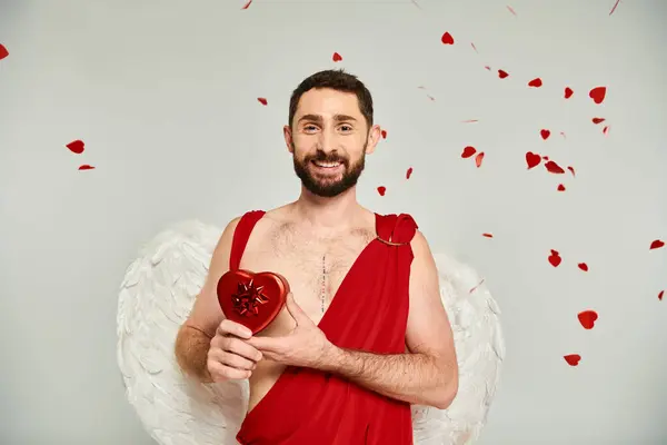 Hombre barbudo feliz vestido como Cupido con San Valentín rojo en forma de oído presente en gris, fiesta de disfraces - foto de stock