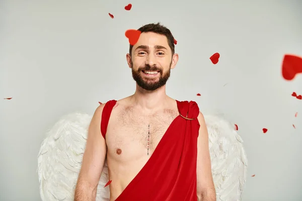 Bärtiger Mann in Amor-Kostüm und Flügeln, der unter rotem herzförmigem Konfetti auf grau in die Kamera lächelt — Stockfoto