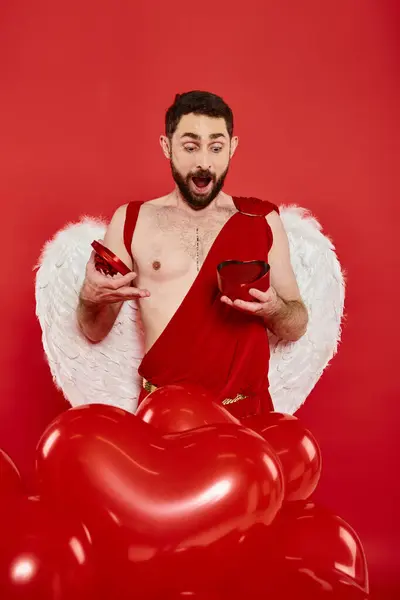 Здивований чоловік в куполоподібному костюмі, що відкриває день святого Валентина, присутній біля повітряних кульок у формі серця на червоному — стокове фото