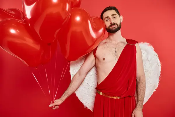 Uomo cupido sognante e felice con palloncini a forma di cuore guardando la fotocamera in rosso, giorno di San Valentino — Foto stock