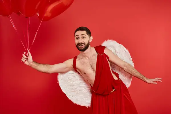 Uomo barbuto sorridente in costume cupido in posa con palloncini a forma di cuore su rosso, San Valentino — Foto stock