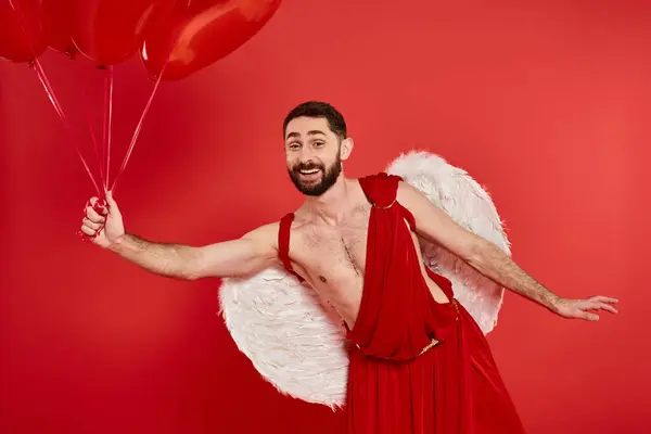 Smiling bearded man in cupid costume posing with heart-shaped balloons on red, st valentines day — Stock Photo