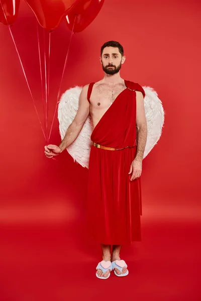 Offended bearded man dressed as cupid with heart-shaped balloons on red, st valentines costume party — Stock Photo