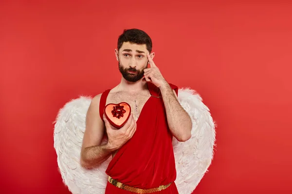 Вдумливий бородатий чоловік у костюмі з серцеподібною подарунковою коробкою, що вказує на голову на червоний — стокове фото