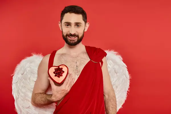 Smiling bearded cupid man showing heart-shaped gift box on red, st valentines day themed event — Stock Photo