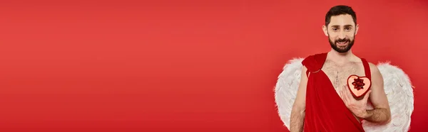 Satisfeito barbudo cupido homem mostrando coração em forma de st valentines presente no vermelho, festa traje, banner — Fotografia de Stock