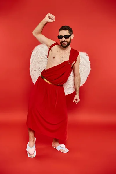 Pleine longueur de l'homme excité en costume de Cupidon et lunettes de soleil dansant sur le rouge, st fête de la Saint-Valentin — Photo de stock