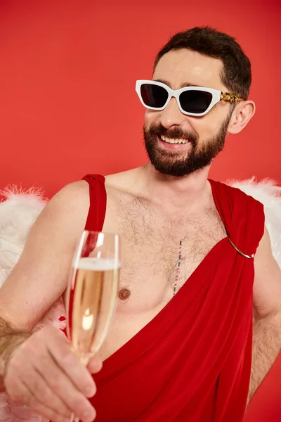 Homme barbu en lunettes de soleil et costume de Cupidon souriant avec verre de champagne en rouge, st Saint-Valentin — Photo de stock