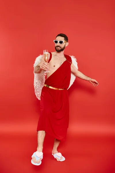 Повна довжина грайливого чоловіка в сонцезахисних окулярах і куполоподібному костюмі позує з шампанським на червоному — стокове фото