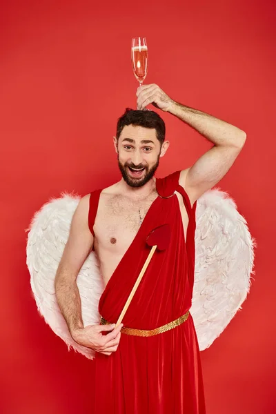 Hombre divertido y alegre en traje de Cupido posando con flecha en forma de corazón y copa de champán en la cabeza - foto de stock