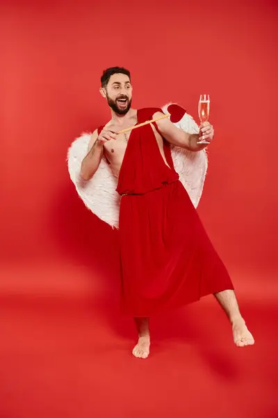 Uomo barbuto eccitato in costume cupido con freccia a forma di cuore e bicchiere di champagne su rosso, lunghezza intera — Foto stock
