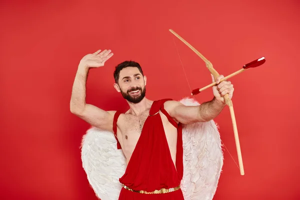Cheerful man in cupid costume archering with heart-shaped arrow on red, st valentines costume party — Stock Photo
