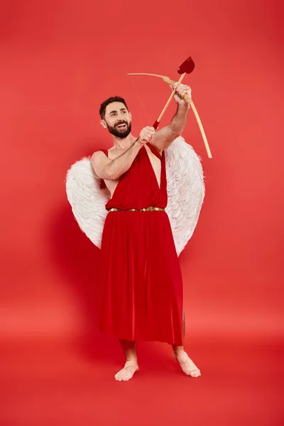 Piena lunghezza di uomo eccitato in costume cupido tiro con l'arco sul rosso, concetto di San Valentino — Foto stock
