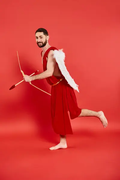 Drôle barbu homme en costume de Cupidon courir avec arc et flèche sur rouge, st Saint Valentin célébration — Photo de stock