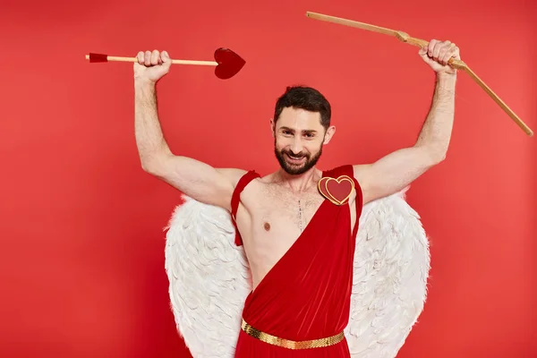 Freudiger bärtiger Mann im Amor-Kostüm mit Bogen und herzförmigen Pfeilen in erhobenen Händen auf rot — Stockfoto