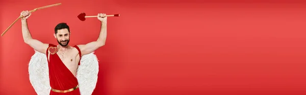 Homme cupide barbu énergisé avec arc et flèche en forme de coeur dans les mains levées sur rouge, bannière — Photo de stock