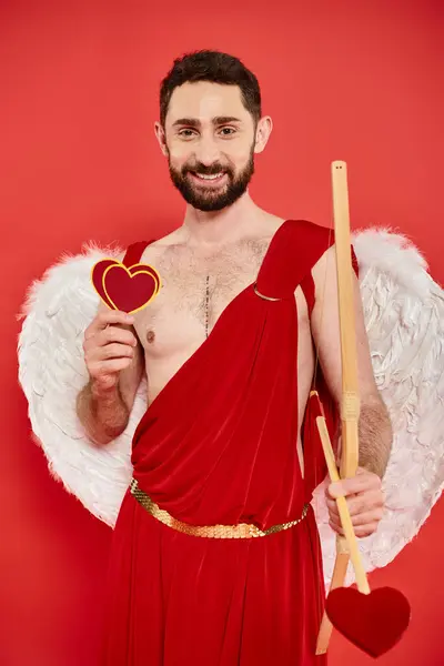 Glücklicher bärtiger Mann im Amor-Kostüm mit Papierherzen und Bogen mit Pfeil am roten Valentinstag — Stockfoto