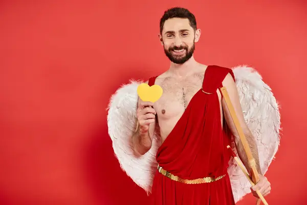 Allegro uomo barbuto in costume cupido con fiocco e cuore di carta gialla su rosso, San Valentino — Foto stock