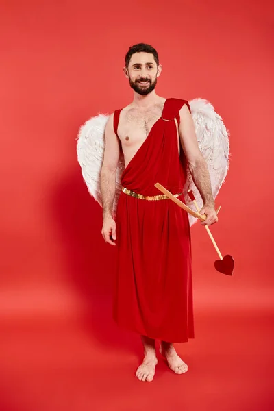 Uomo barbuto sorridente a piedi nudi in costume cupido con arco e freccia in piedi e guardando lontano sul rosso — Foto stock