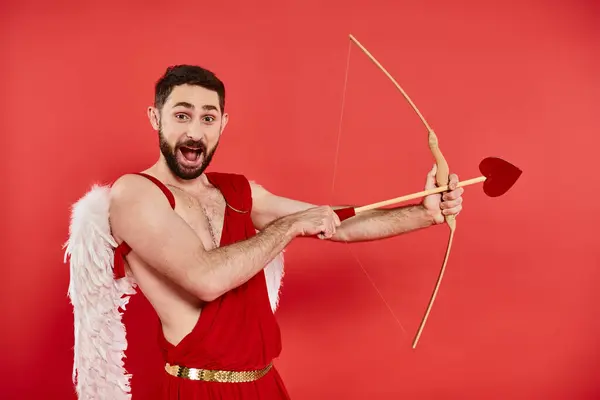 Excited cupid man with open mouth archering and looking at camera on red, st valentines concept — Stock Photo