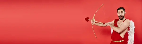 Thrilled cupid man with open mouth archering while looking at camera on red, horizontal banner — Stock Photo