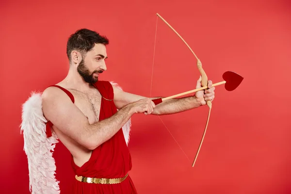 Vista laterale di uomo barbuto in costume cupido mira e tiro con l'arco con freccia a forma di sentire sul rosso — Foto stock