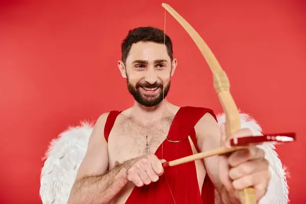 Joyeux homme barbu en costume de Cupidon regardant loin tout en visant et tir à l'arc sur rouge, st valentines — Photo de stock