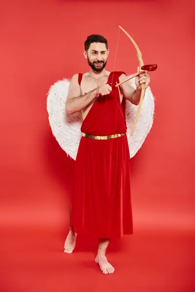 Full length of smiling barefoot man in cupid costume looking away and archering on red backdrop — Stock Photo