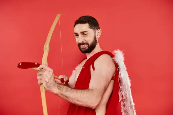 Щасливий бородатий чоловік, одягнений як купідон стрільба зі стрілою у формі серця на червоному, день Святого Валентина — стокове фото
