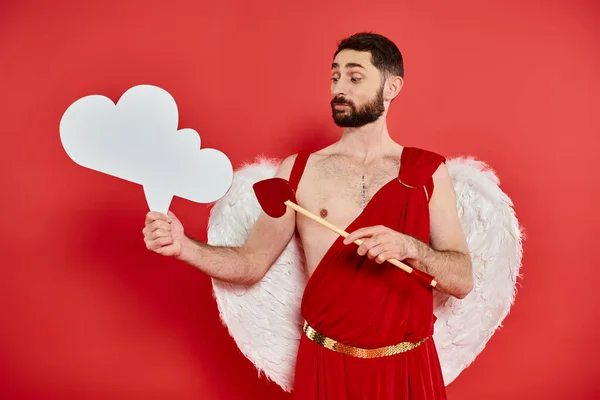 Pensive cupid man with heart-shaped arrow looking at white empty thought-bubble on red backdrop — Stock Photo