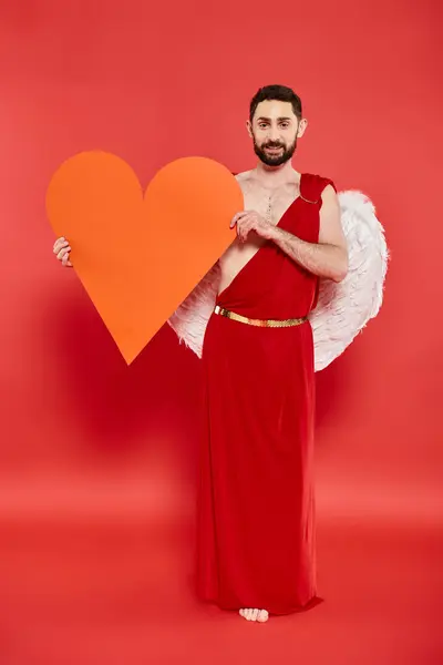 Full length of bearded man dressed as cupid with huge paper heart on red, valentines costume party — Stock Photo