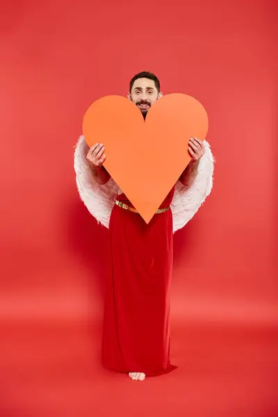 Smiling bearded man dressed as cupid holding huge orange heart on red, valentines costume party — Stock Photo