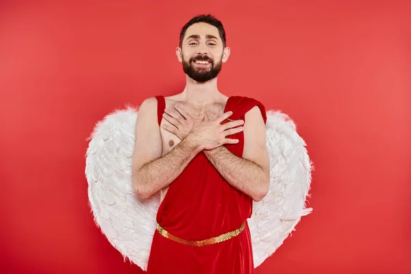 Hombre barbudo feliz en traje de Cupido de pie con las manos en el pecho y mirando a la cámara en rojo - foto de stock