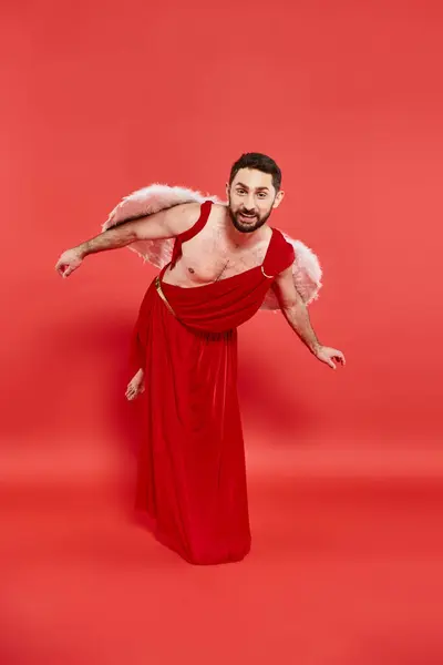 Engraçado barbudo homem vestido como cupido curvando e olhando para a câmera em vermelho, st valentines evento temático — Fotografia de Stock
