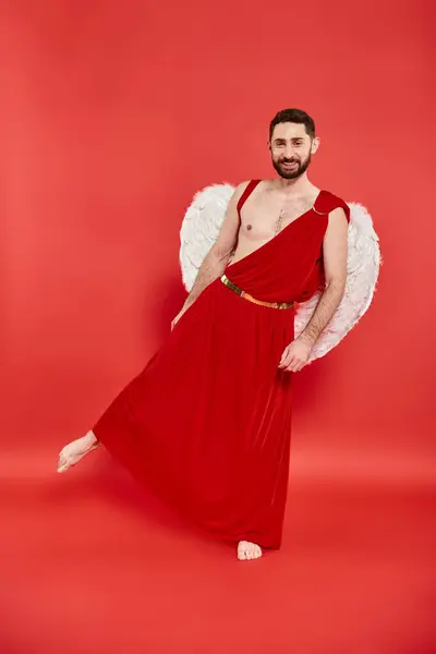 Homme barbu en costume de Cupidon et les ailes debout dans la pose théâtrale sur le rouge, Saint Valentin — Photo de stock